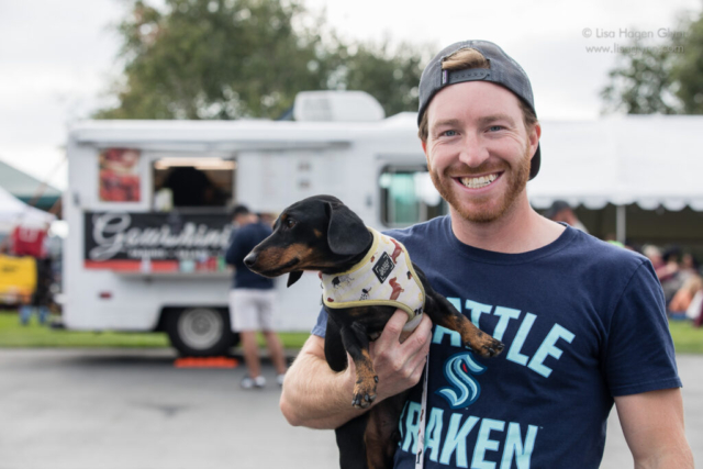 A person holds a wiener dog wearing a wiener-dog themed harness, with a food truck in the distance
