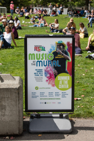 A sign advertises the Music at the Mural series. An audience sits in the grass behind.