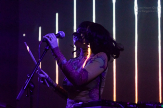 Kelly Lee Owens flings her hair to the right while holding a microphone in her left hand and a drumstick in her right