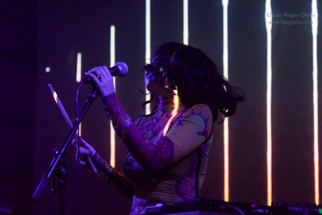 Kelly Lee Owens flings her hair to the right while holding a microphone in her left hand and a drumstick in her right