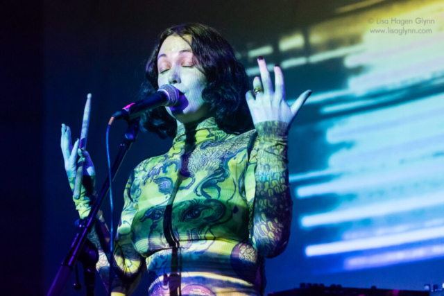 Kelly Lee Owens closes her eyes while singing and holding her forearms in the air