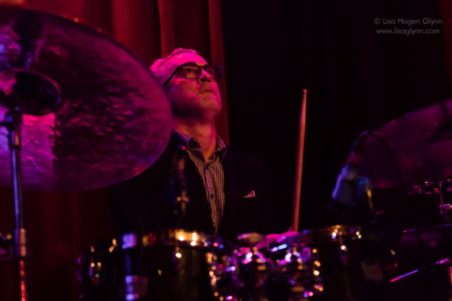 A person wearing glasses looks upward while playing drums