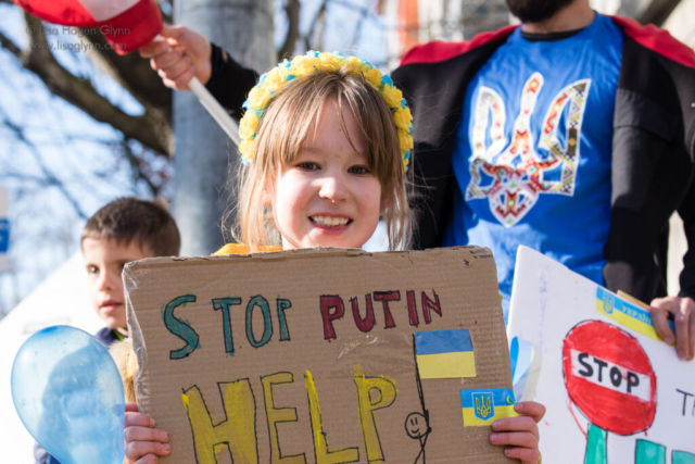 A child wears a yellow and blue floral headdress and carries a sign that reads, "Stop Putin. Help."