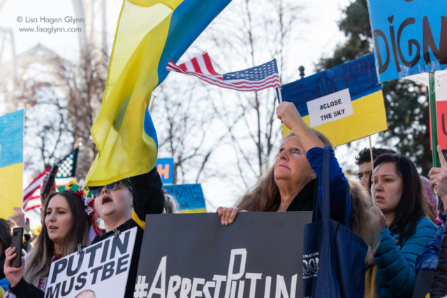 A person waves a United States flag, while holding a sign that reads "#ArrestPutin"